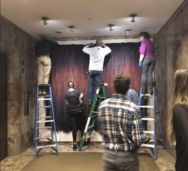 Several museum staff members standing on ladders adjust and install a large dark red and black textile wall hanging in a gallery space.